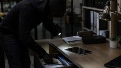 Vermummter Einbrecher mit Handschuhen und Taschenlampe entwendet Bargeld aus Schreibtischschublade in einem Büro