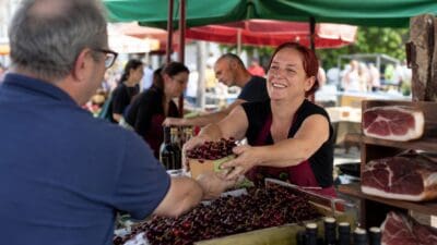 Eine lächelnde Marktfrau mit roten Haaren übergibt einem Mann einen Korb mit Kirschen.