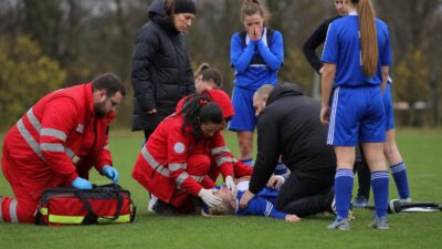 Rettungskräfte in roten Jacken behandeln eine am Boden liegende Fußballspielerin auf grünem Rasen.