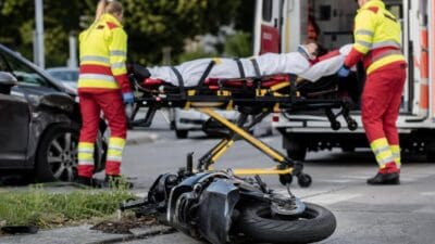Sanitäter beladen einen Rettungswagen mit einem Patienten auf einer Trage an einer Unfallstelle mit umgestürztem Moped.