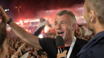 Ein grauhaariger Mann singt ins Mikrofon und hebt den Arm vor einer jubelnden Menschenmenge bei Nacht.