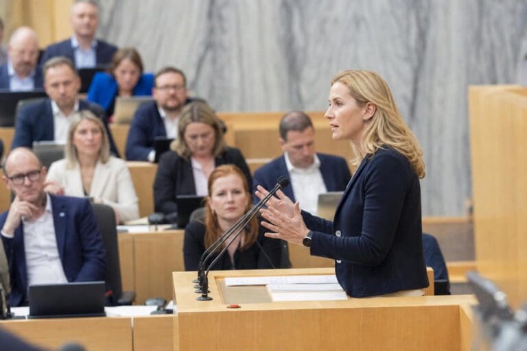Blonde Politikerin gestikuliert während einer Rede am Pult im österreichischen Nationalrat vor sitzenden Abgeordneten