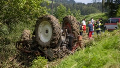 Umgestürzter Traktor mit großen Rädern liegt in einer Wiese am Waldrand