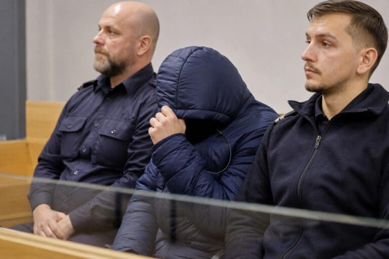 Drei Männer sitzen im Gerichtssaal, einer verdeckt mit hochgezogener Kapuze sein Gesicht.