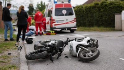 Umgestürztes weißes Moped liegt auf der Straße, Rettungskräfte versorgen Verletzte neben einem Krankenwagen.