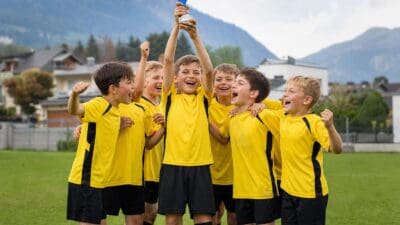 Junge Fußballspieler in gelben Trikots halten einen Pokal hoch und jubeln auf einem grünen Spielfeld.