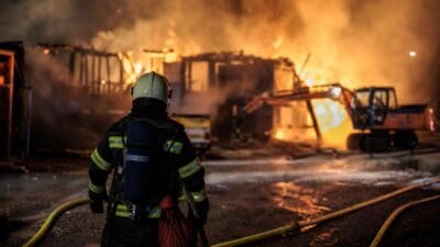 Rückenansicht eines Feuerwehrmanns vor brennendem Gebäude mit Bagger und Schläuchen bei Nacht