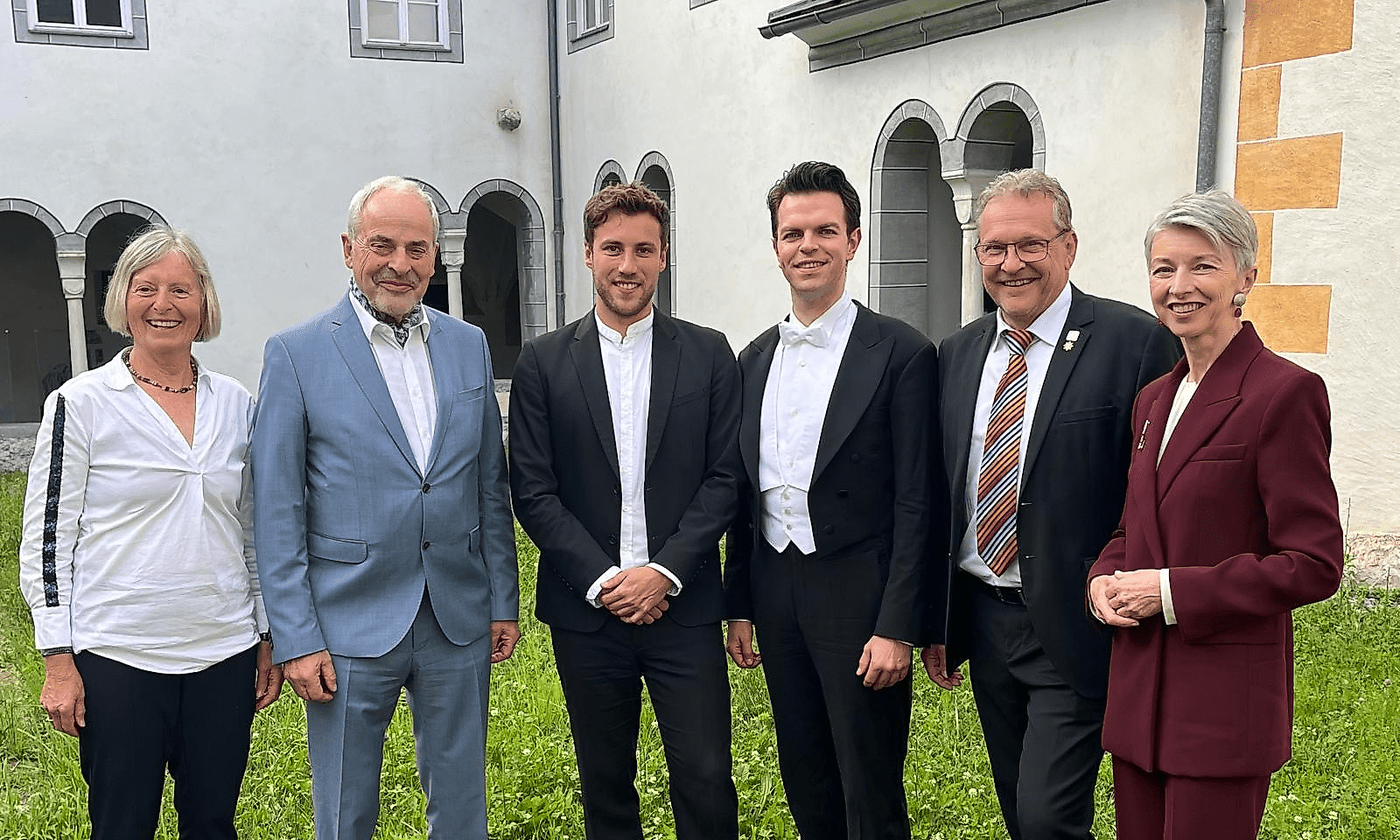 Sechs Personen in formeller Kleidung stehen auf einer Wiese vor einem Gebäude mit Arkaden.