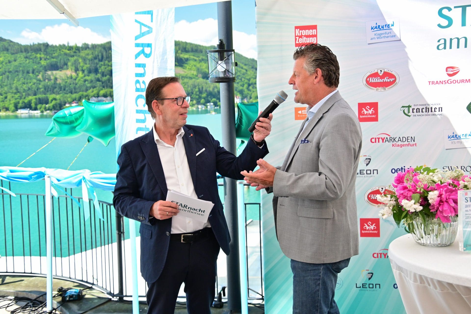 Zwei Männer in Anzügen bei einer Veranstaltung am Wörthersee, einer spricht ins Mikrofon.