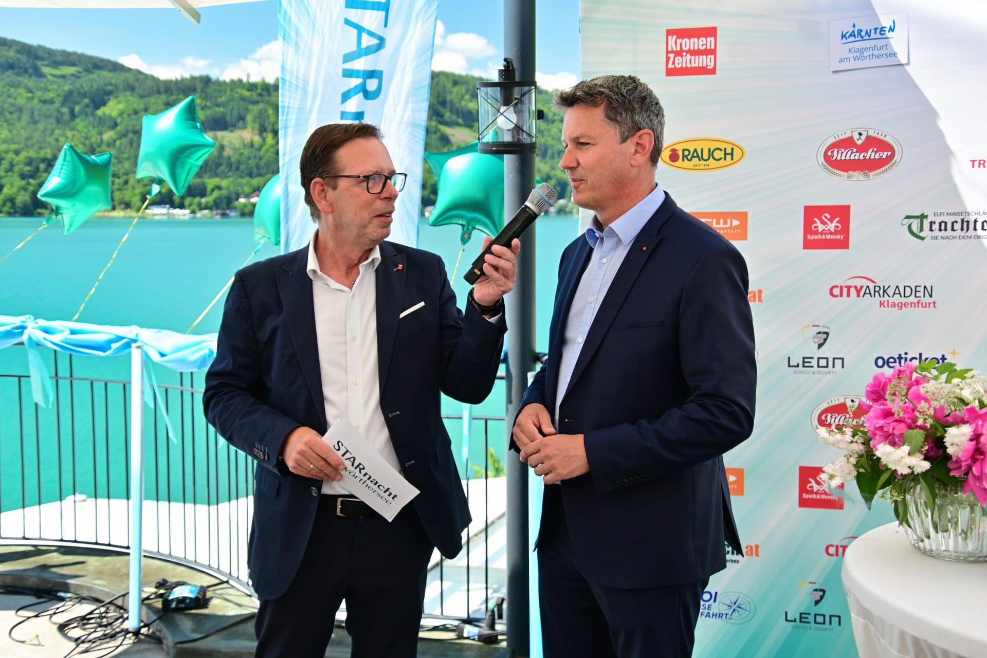 Zwei Männer in dunklen Anzügen auf einer Bühne am Wörthersee, einer hält ein Mikrofon.