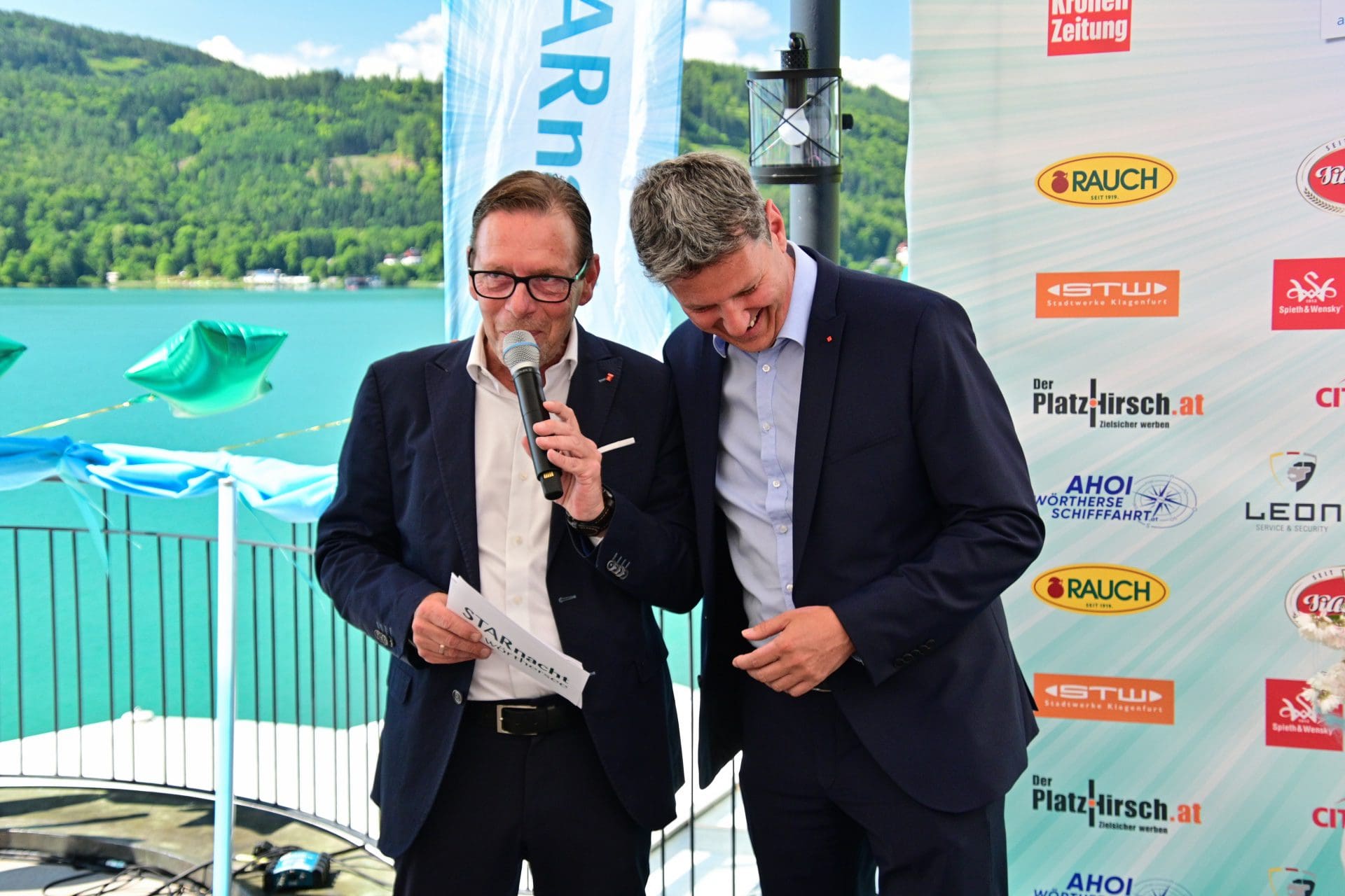 Zwei Männer in Anzügen bei einer Veranstaltung am Wörthersee, einer spricht ins Mikrofon.
