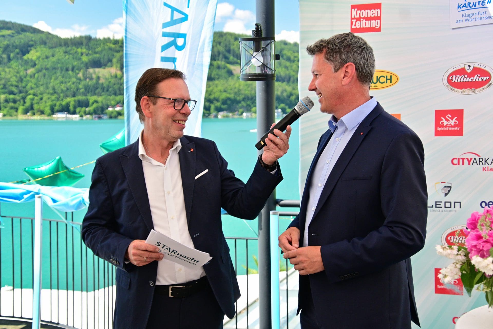 Zwei Männer in Anzügen auf einer Bühne am Wörthersee, einer hält ein Mikrofon