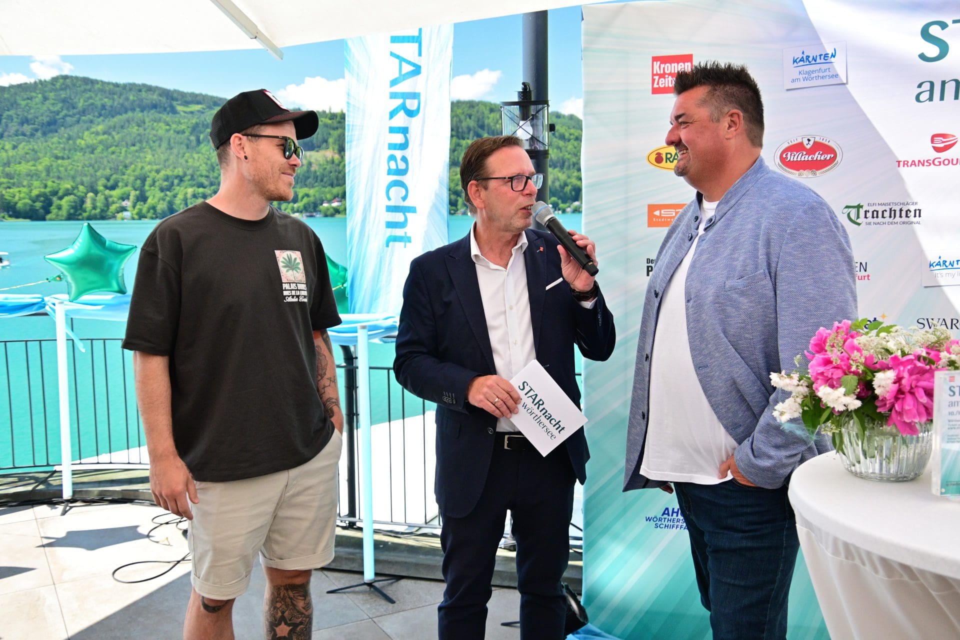 Drei Männer bei einer Pressekonferenz am Ufer des Wörthersees mit Sponsorenbannern im Hintergrund