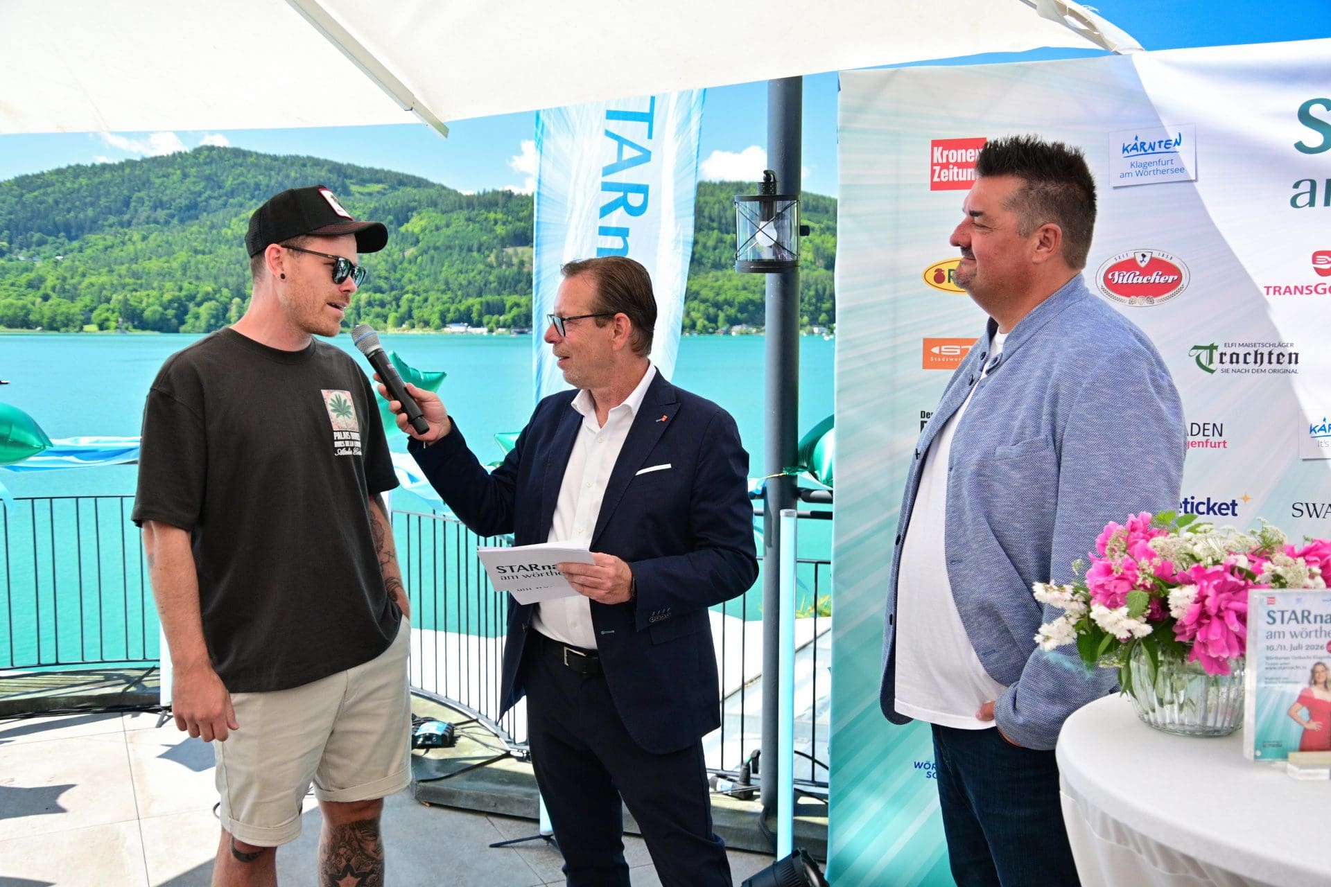 Drei Männer bei einer Pressekonferenz am Wörthersee mit Mikrofon und Bannern