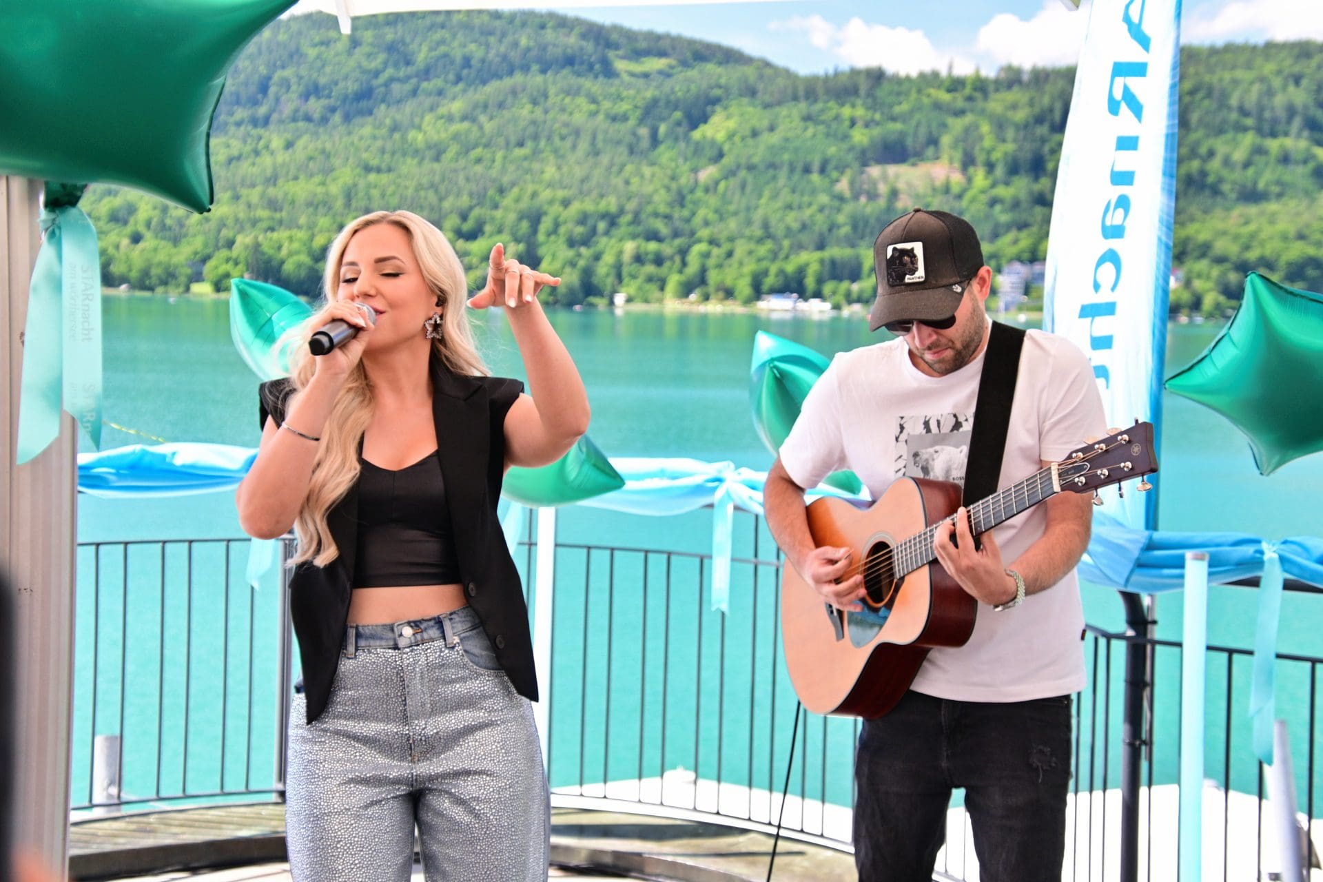 Blonde Sängerin singt in ein Mikrofon während ein Gitarrist auf der Bühne spielt