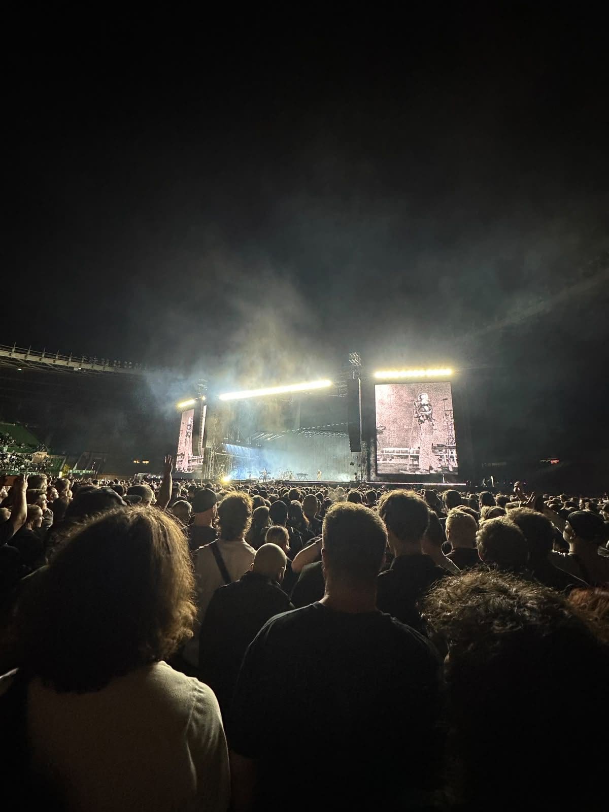 Blick von hinten auf eine dichte Menschenmenge bei einem nächtlichen Konzert in einem Stadion mit beleuchteter Bühne.