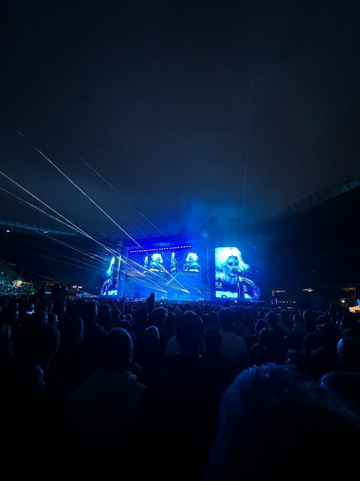 Dichte Menschenmenge vor einer blau beleuchteten Konzertbühne mit großen Videoleinwänden und Lasershow