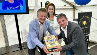 Zwei Männer präsentieren eine Torte bei einer Philips Jubiläumsfeier, eine Frau steht im Hintergrund.