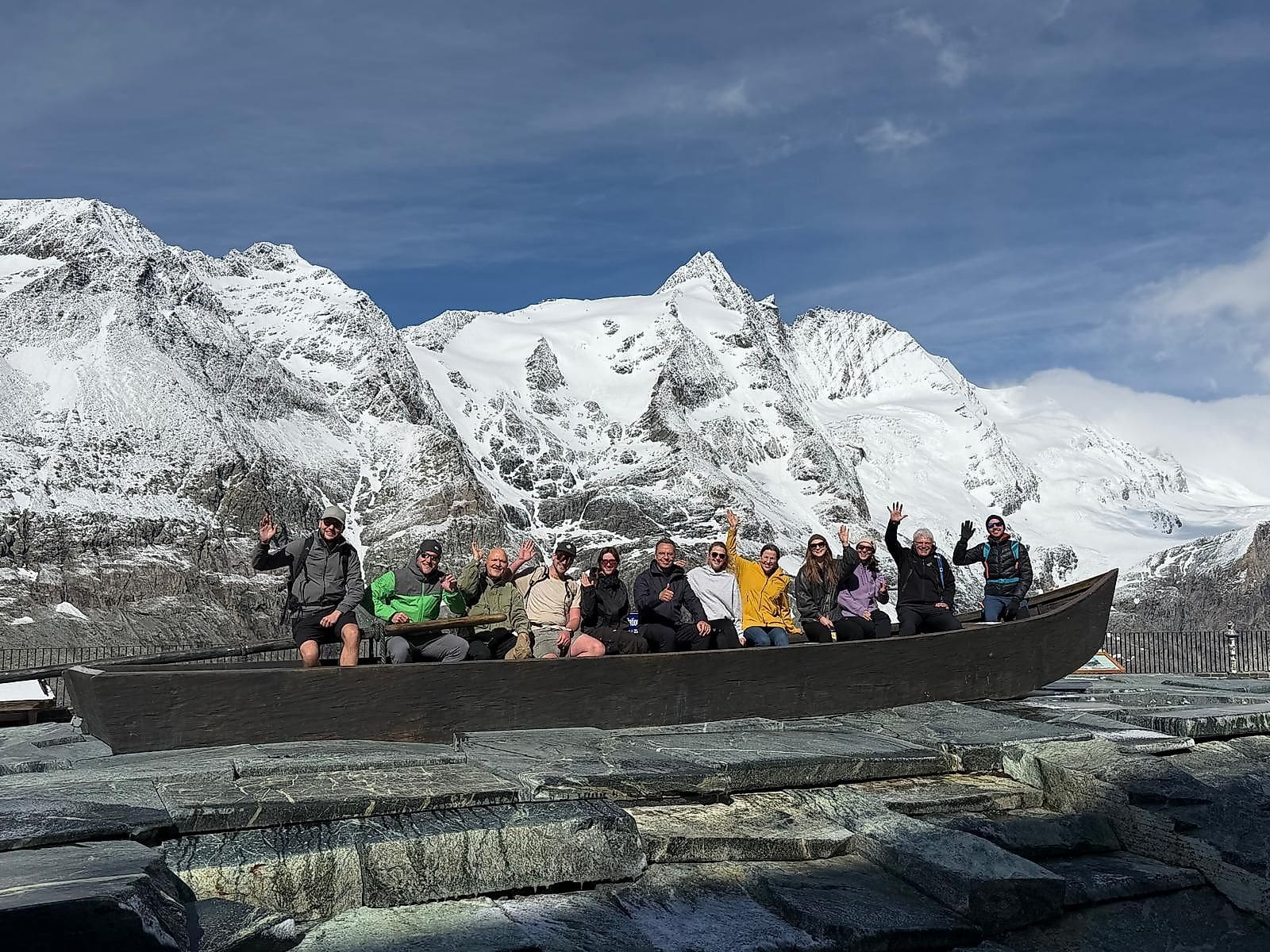 Gruppe von Personen winkt aus einem hölzernen Boot vor schneebedeckten Bergen bei Heiligenblut.