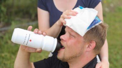 Ein Mann trinkt aus einer Flasche während eine Frau ihm einen kühlen Umschlag auf den Kopf legt.