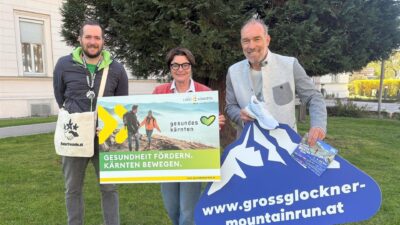 Drei Personen halten Werbeschilder für den Großglockner Mountain Walk auf einer Wiese.