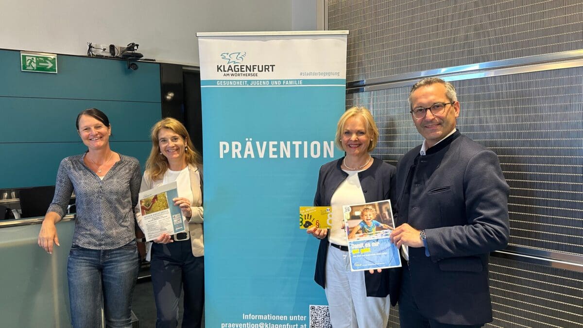 Vier Personen stehen vor einem Präventions-Banner der Stadt Klagenfurt und halten Informationsmaterialien in den Händen.
