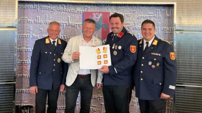 Vier Männer stehen nebeneinander vor einer Wand mit Wappen, drei in Feuerwehruniform und einer hält eine Tafel mit Abzeichen.