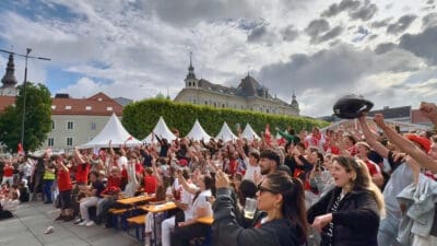 Große Menschenmenge jubelt mit erhobenen Armen beim Public Viewing auf dem Neuen Platz in Klagenfurt.