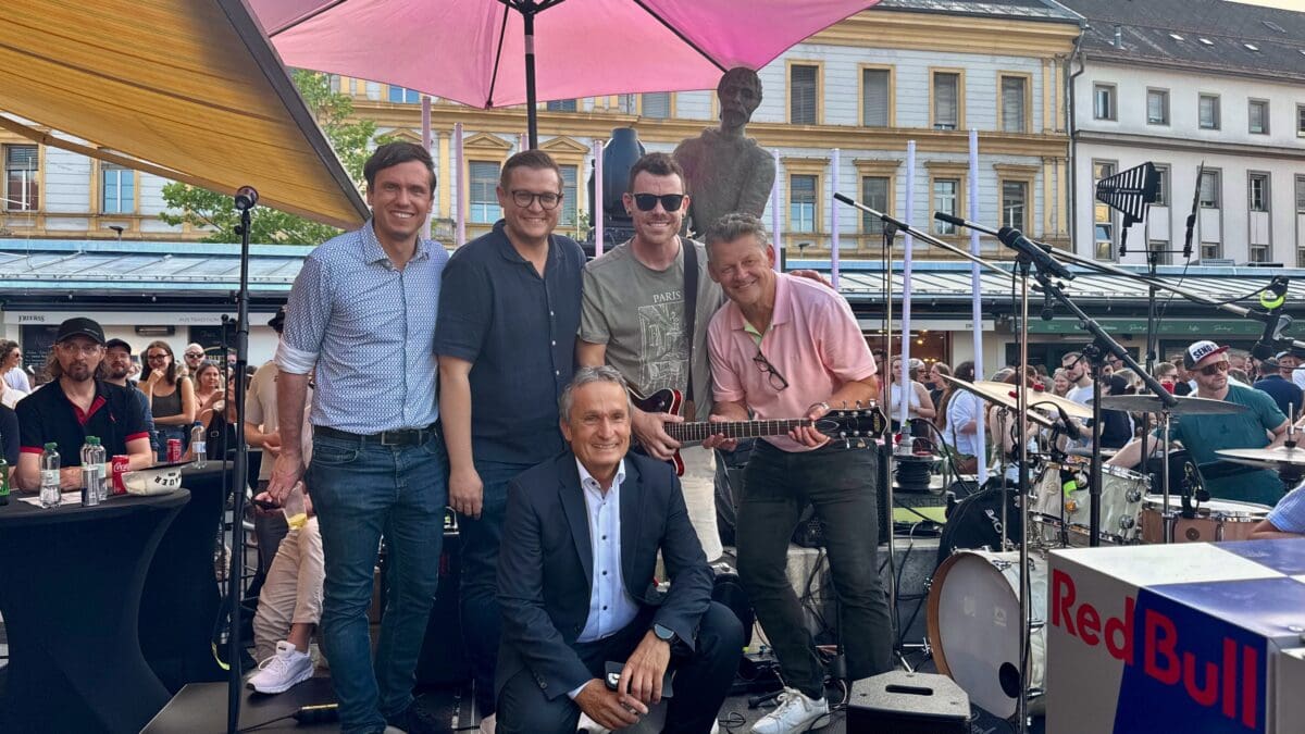Fünf Männer posieren auf einer Bühne mit Musikinstrumenten und pinkem Sonnenschirm.