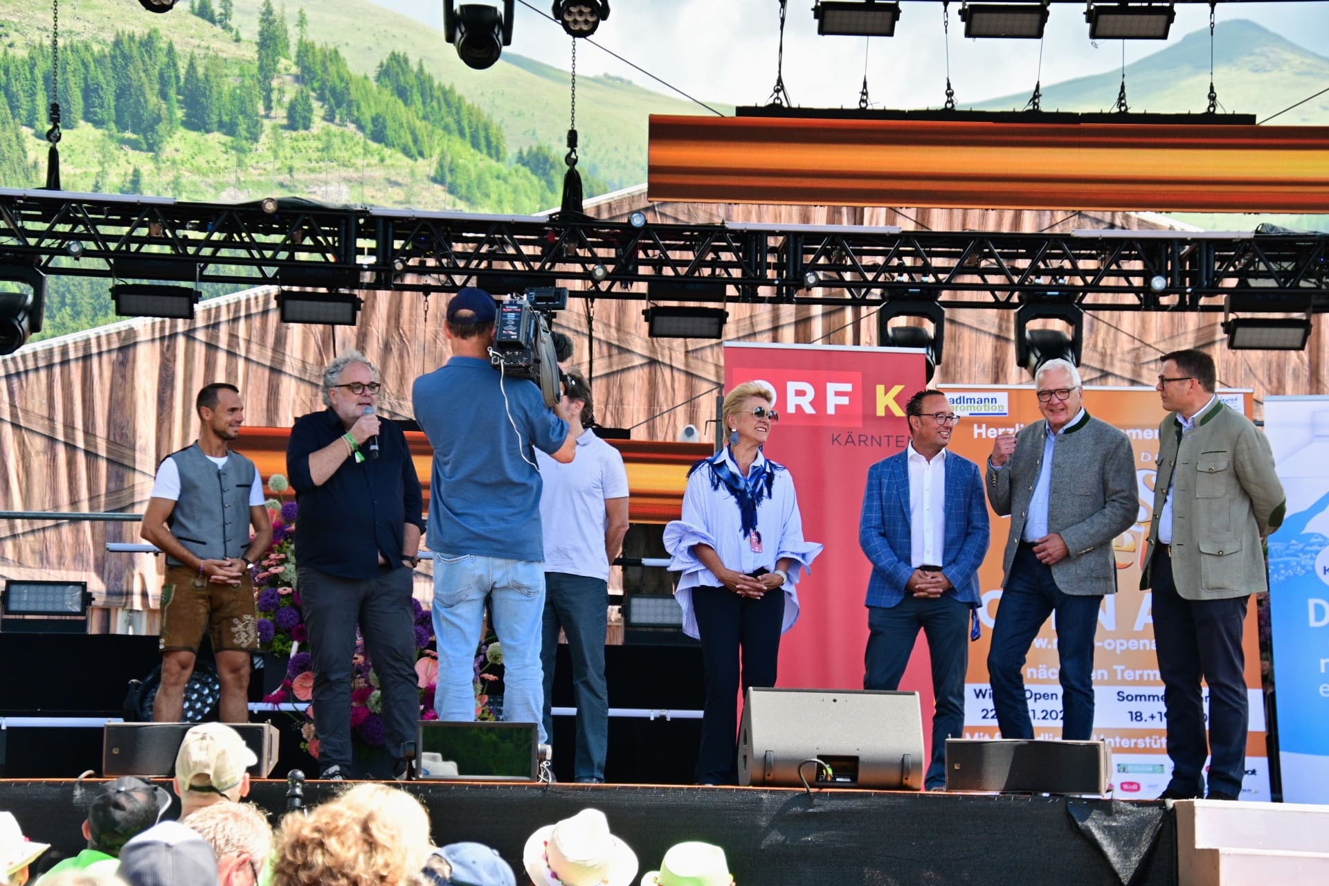 Menschen stehen auf einer Bühne bei einem Open-Air-Event vor Bergpanorama