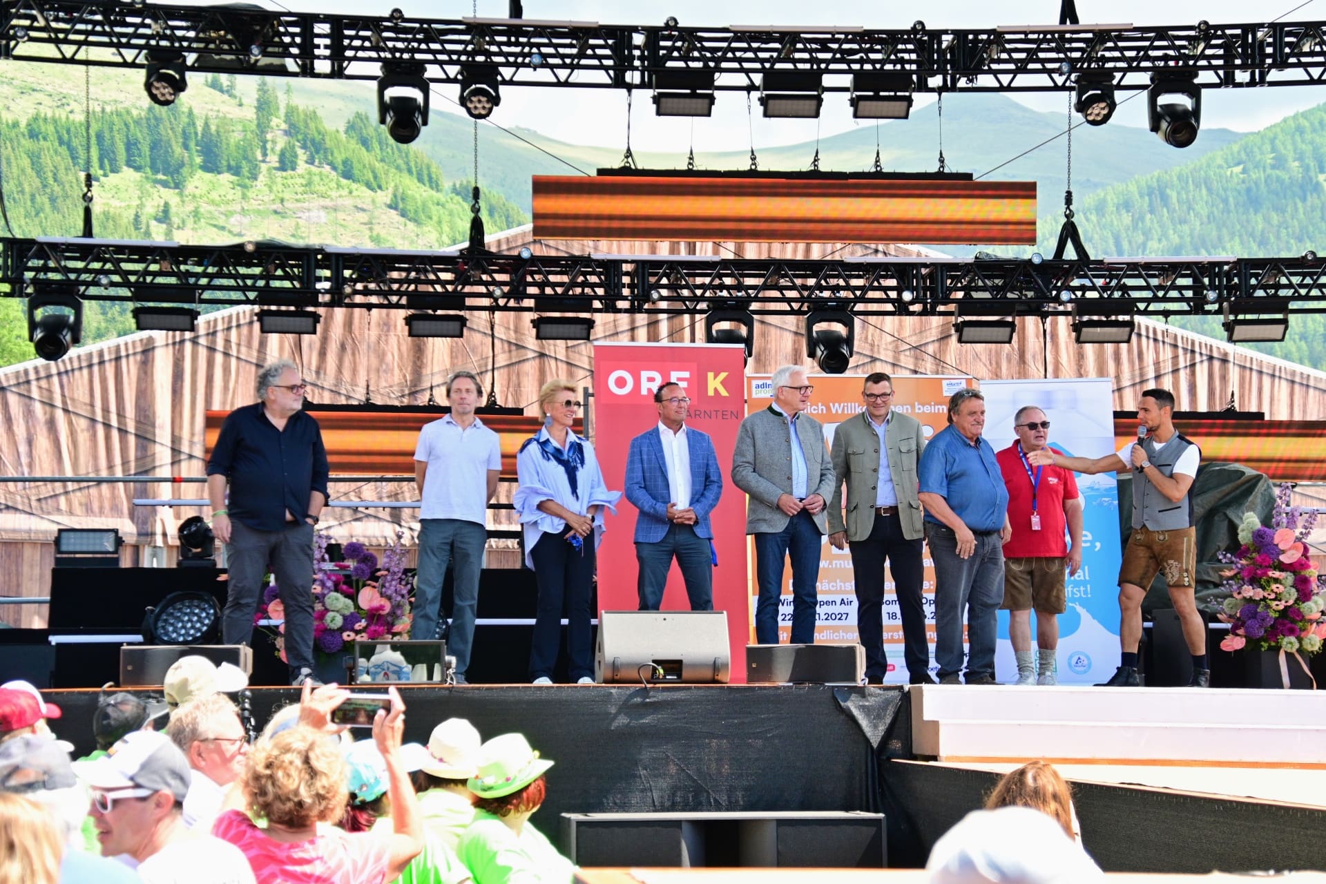 Mehrere Personen stehen auf einer großen Bühne vor einer Bergkulisse bei einem Festival