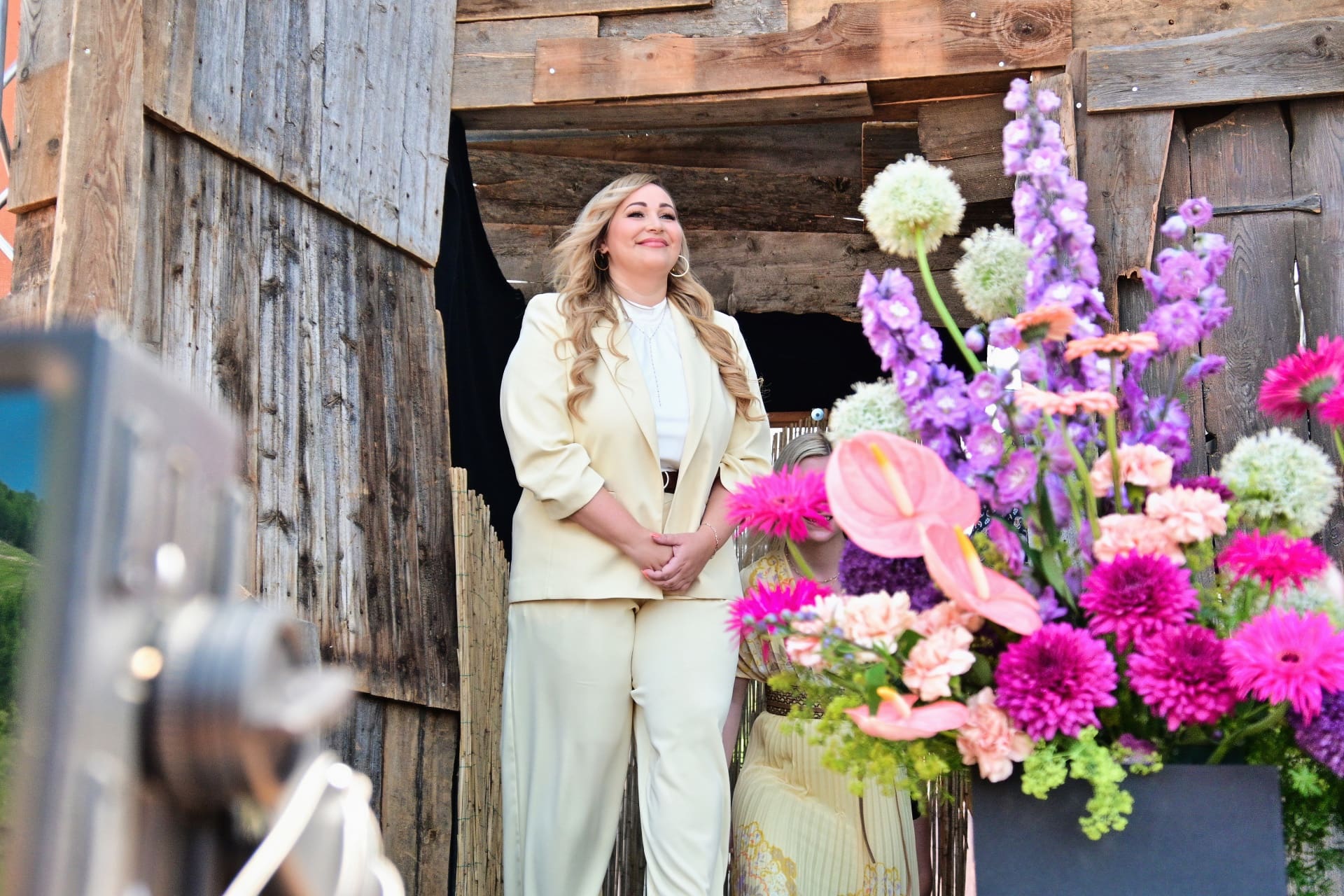 Eine blonde Frau in einem hellgelben Hosenanzug steht lächelnd vor einer Holzkonstruktion mit bunten Blumen im Vordergrund.