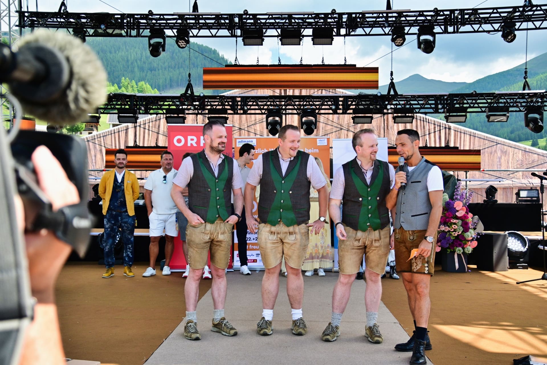 Vier Männer in traditioneller Tracht stehen auf einer Bühne vor Bergen.
