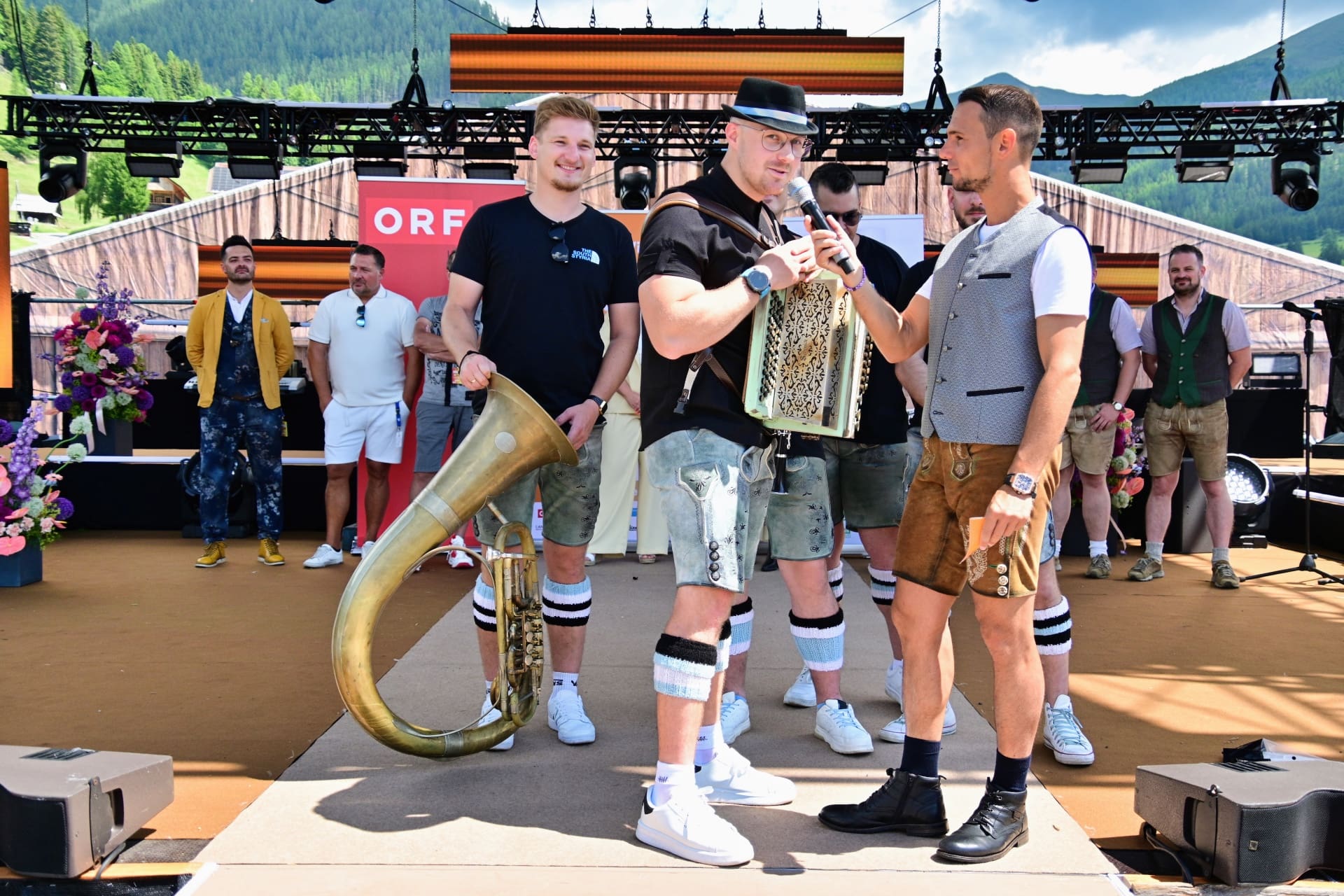 Männer in Tracht mit Akkordeon und Tuba auf einer Open-Air-Bühne vor Bergen.