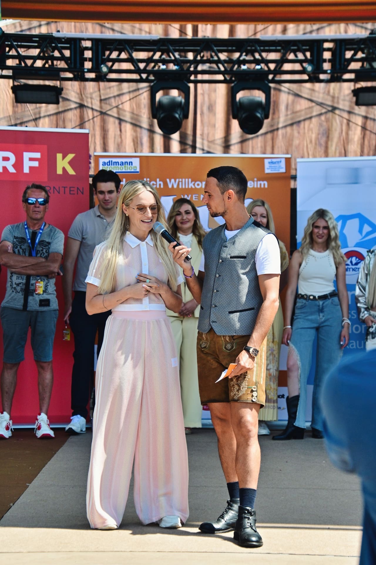 Blonde Frau im rosa Outfit hält Mikrofon und spricht mit Mann in Trachtenweste auf Bühne