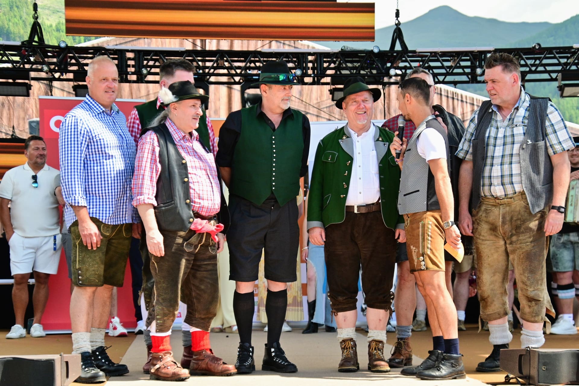 Mehrere Maenner in traditioneller Tracht mit Lederhosen stehen auf einer Buehne.