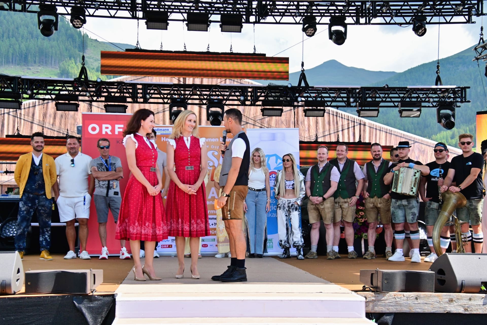 Zwei Frauen in roten Dirndln und ein Moderator mit Mikrofon stehen auf einer Bühne vor Bergkulisse.