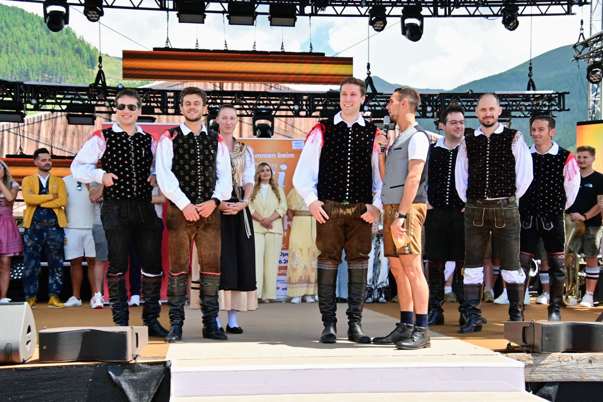 Gruppe von Männern und Frauen in Tracht auf einer Bühne vor Bergkulisse