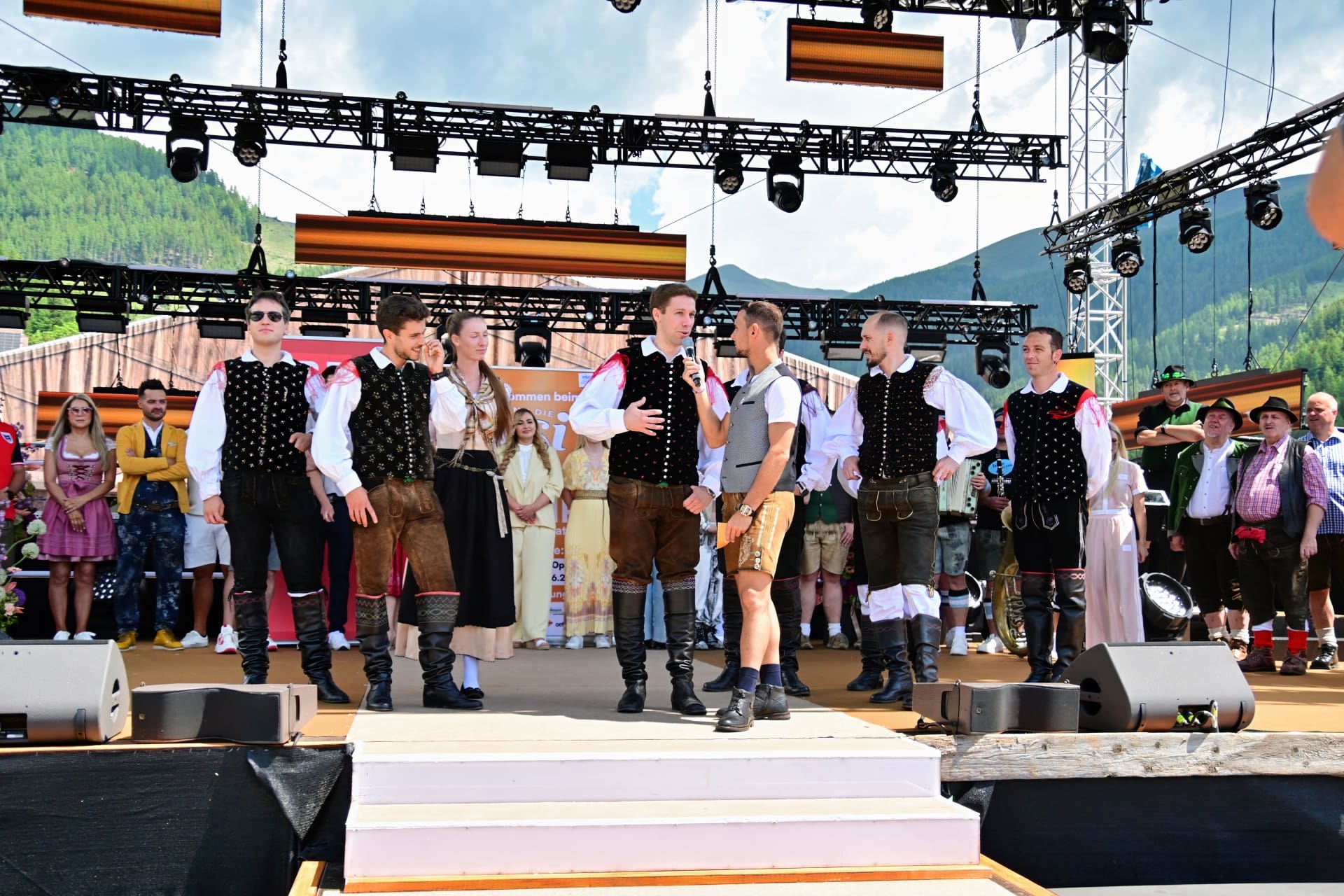 Männer in traditioneller Tracht stehen auf einer Bühne vor Bergkulisse bei einem Open-Air-Event.