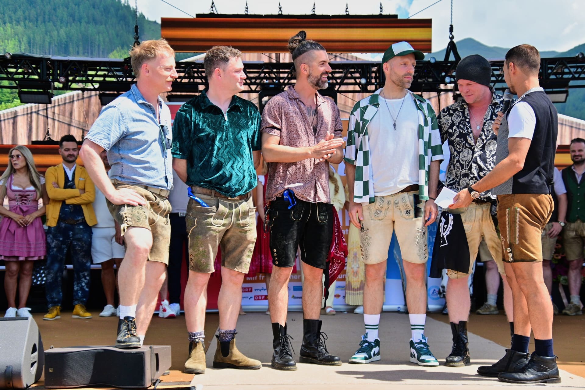 Mehrere Männer in Tracht und moderner Kleidung stehen auf einer Festivalbühne vor Bergkulisse.