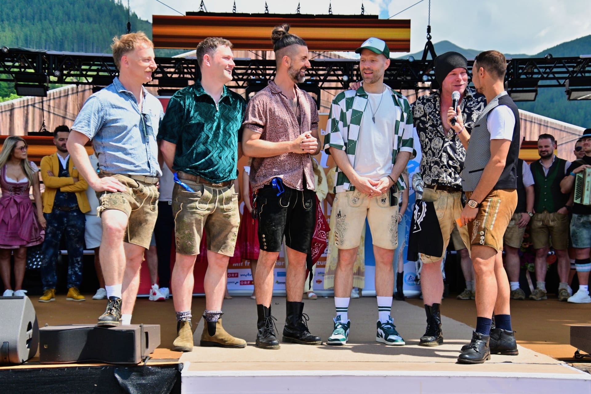 Sechs Männer in Trachtenkleidung stehen auf einer Open-Air-Bühne vor Bergen.
