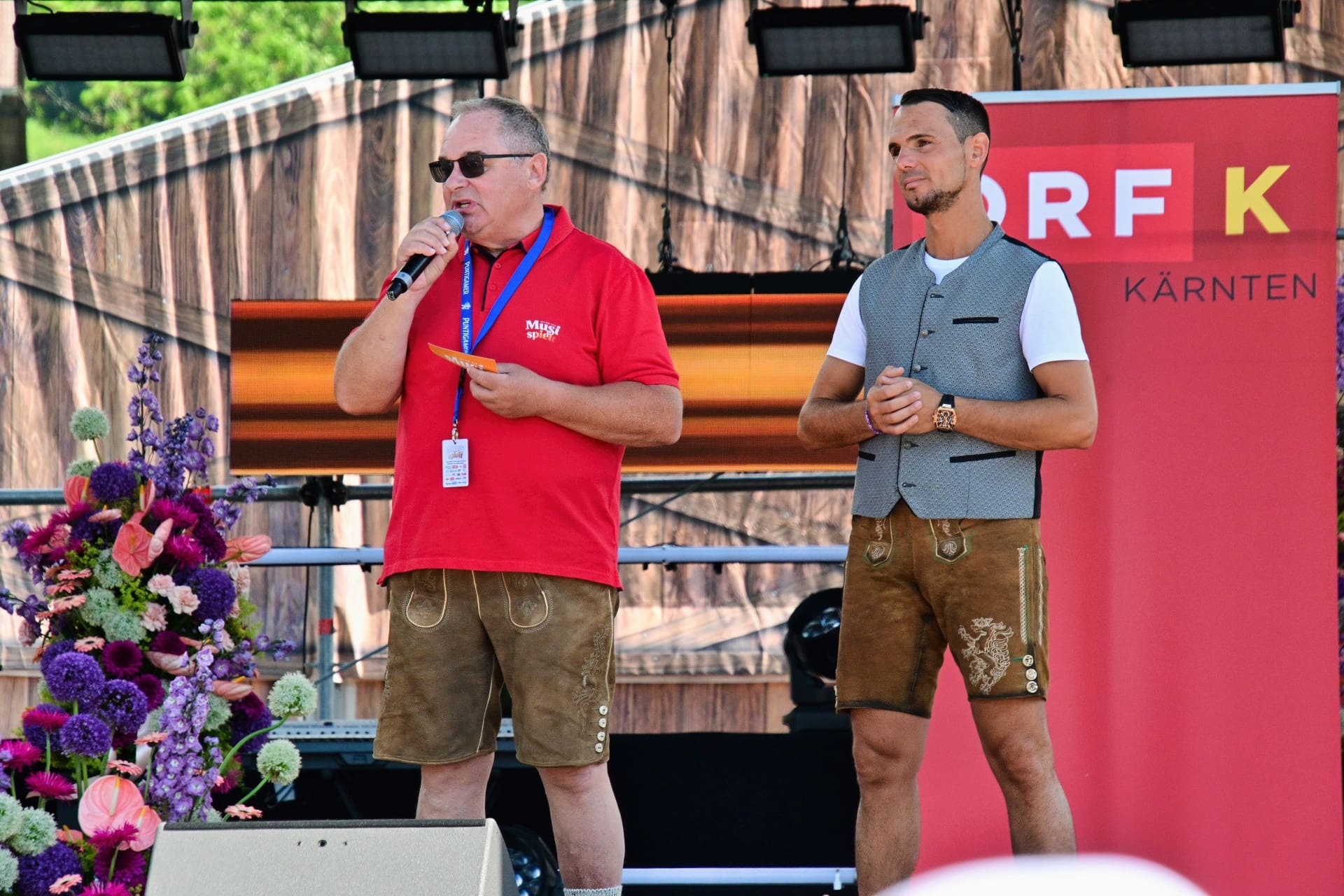 Zwei Männer in Tracht auf einer Bühne, einer spricht ins Mikrofon vor ORF-Banner