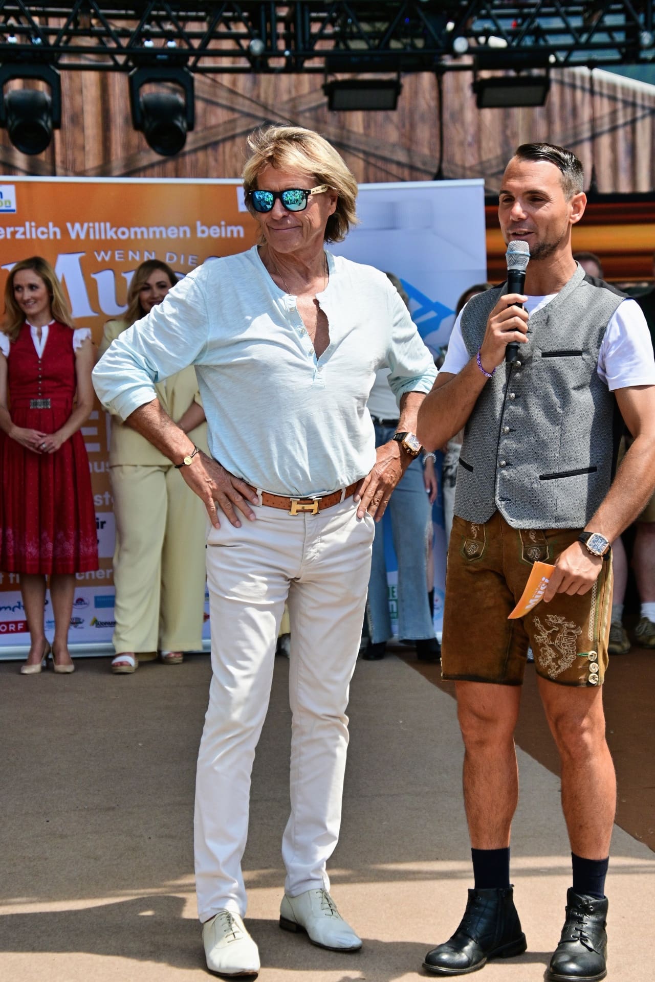 Hansi Hinterseer mit Sonnenbrille in weißer Kleidung neben einem Moderator in Lederhose auf der Bühne.