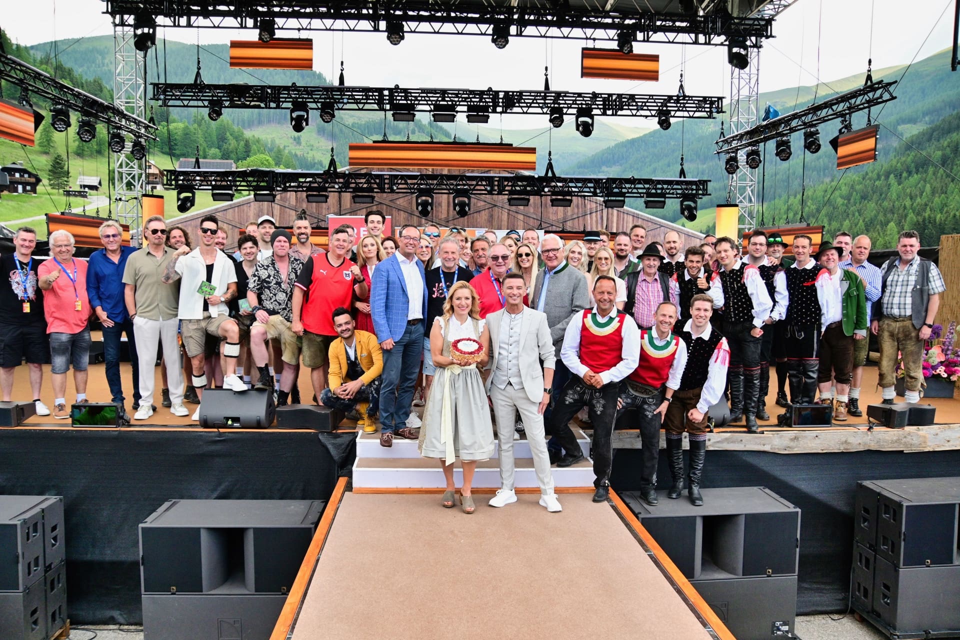 Große Gruppe von Menschen in Tracht und Alltagskleidung posiert auf einer Bühne vor Bergkulisse.