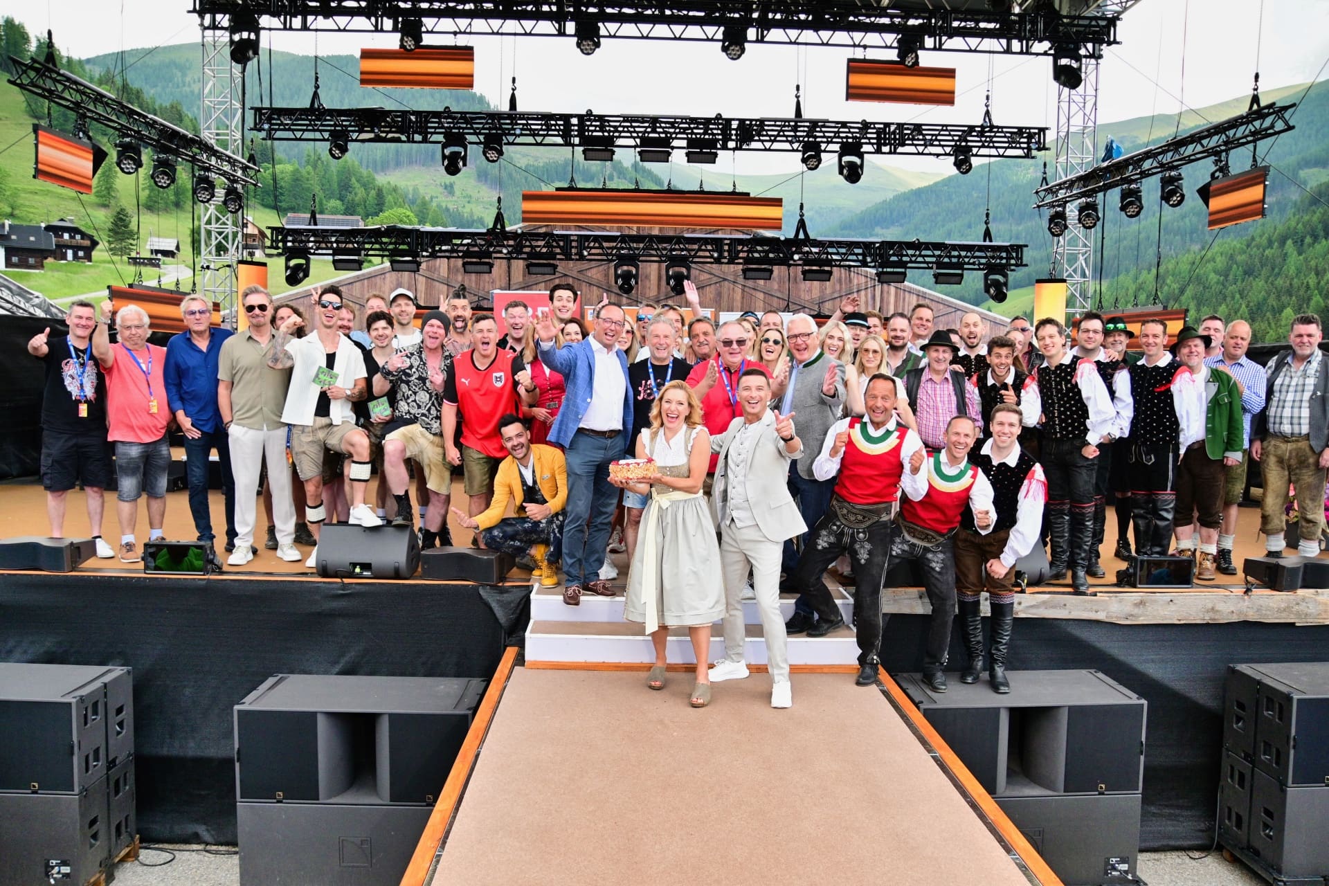 Große Gruppe von Menschen in Tracht und Alltagskleidung posiert gemeinsam auf einer Bühne vor Bergen.