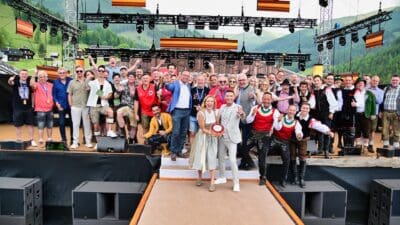 Eine große Gruppe von Menschen in traditioneller und moderner Kleidung steht fröhlich auf einer Bühne vor einer Bergkulisse.