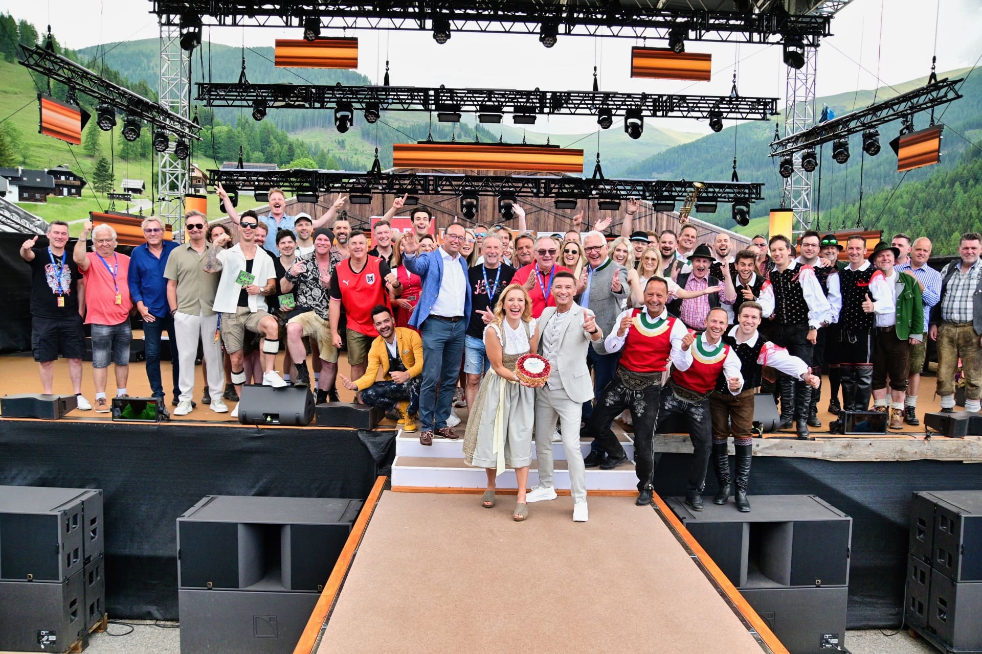 Eine große Gruppe von Menschen in traditioneller und moderner Kleidung steht fröhlich auf einer Bühne vor einer Bergkulisse.