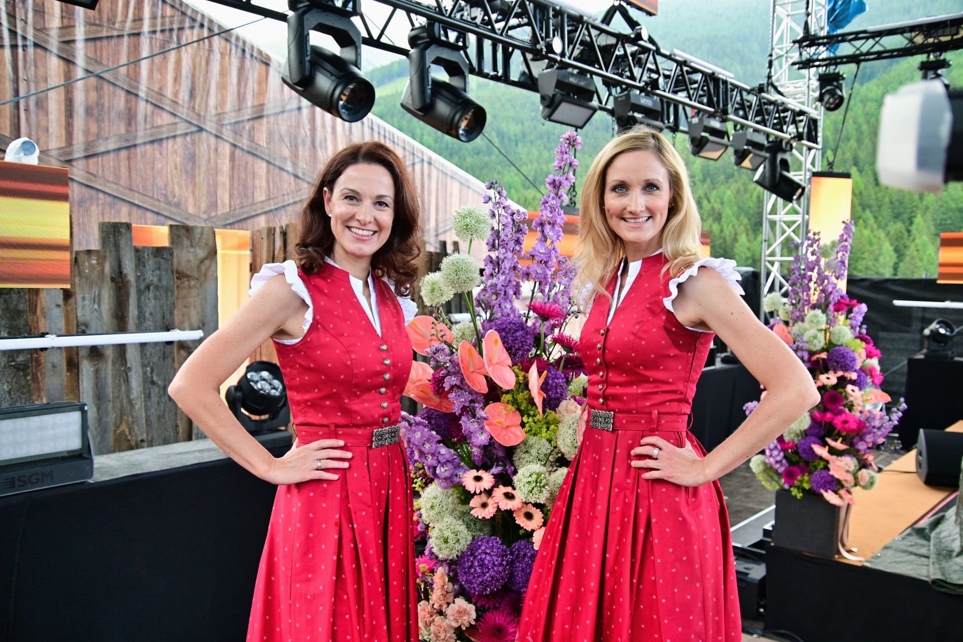 Zwei Frauen in roten Kleidern mit weißen Details stehen neben einem großen bunten Blumenarrangement auf einer Bühne.