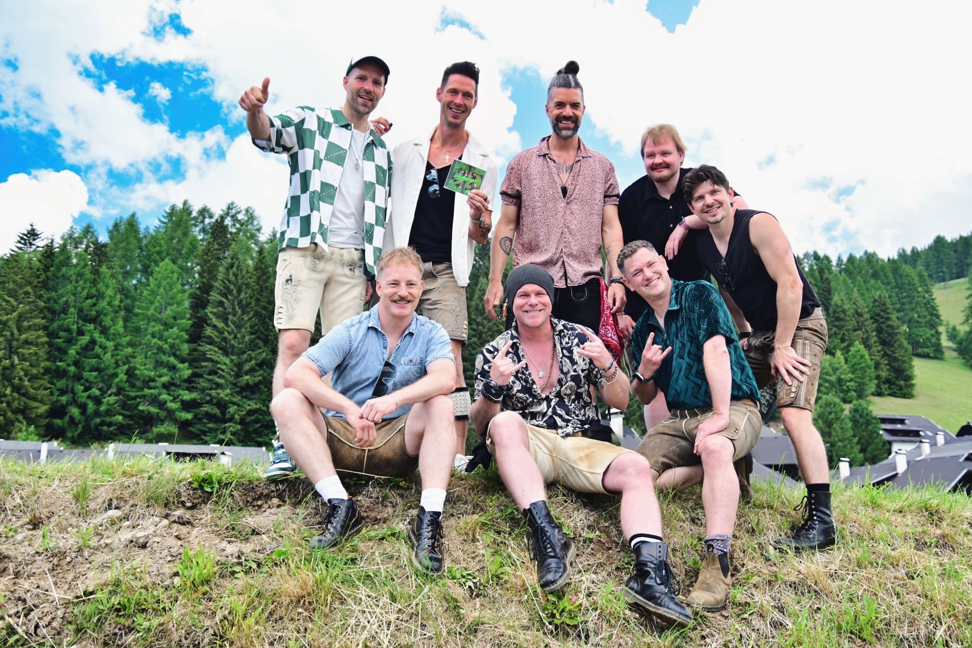 Acht Männer in bunten Hemden und Lederhosen posieren lachend auf einer Wiese vor Bergen.