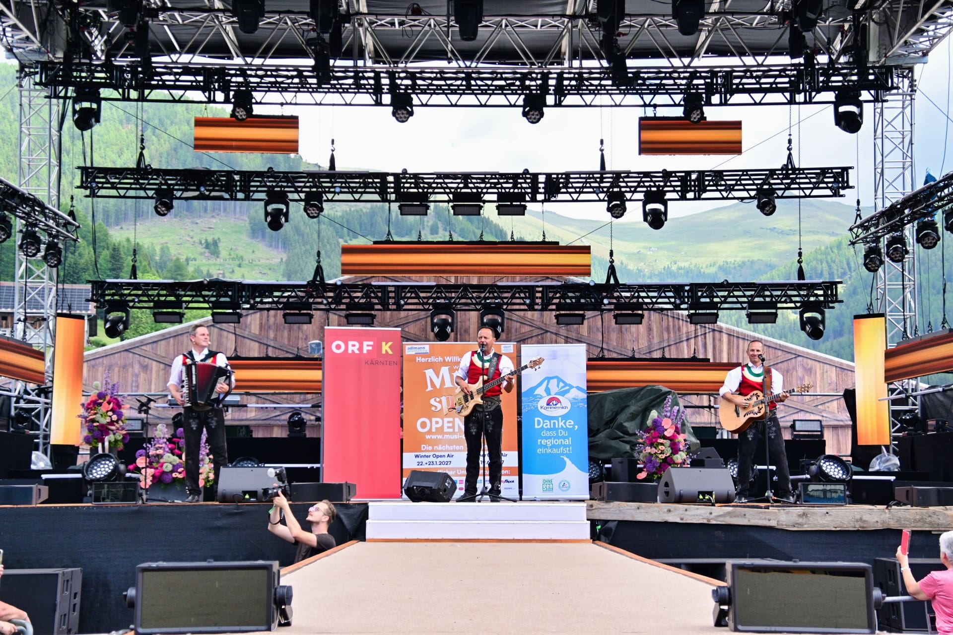 Drei Musiker in Tracht auf einer Open-Air-Bühne mit Akkordeon, Bassgitarre und Akustikgitarre.
